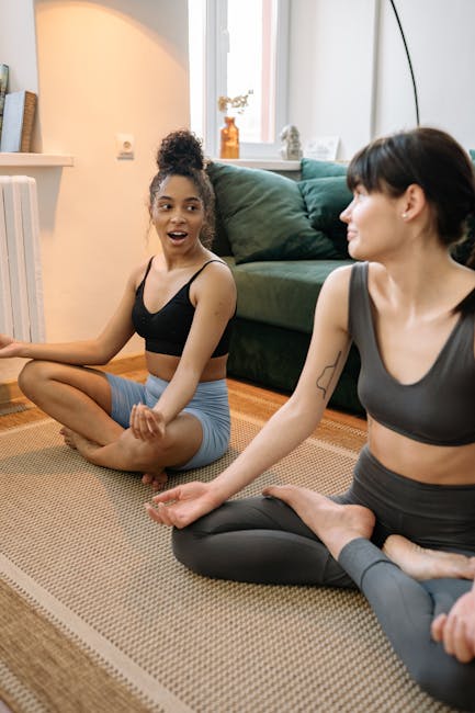 Two women in active wear practicing yoga indoors, sitting cross-legged and smiling.